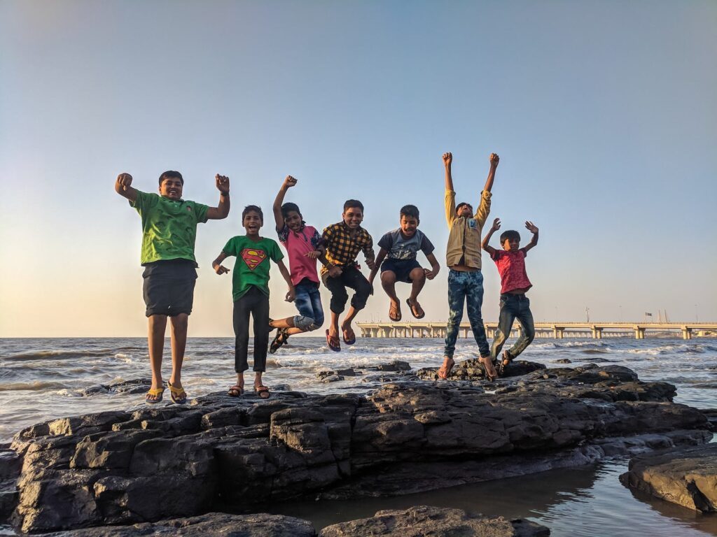 Boy Wearing Green Crew-neck Shirt Jumping from Black Stone on Seashore. self-esteem